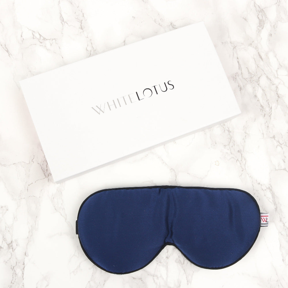 A navy silk eye mask on a marble lifestyle background with an image of the packaging box